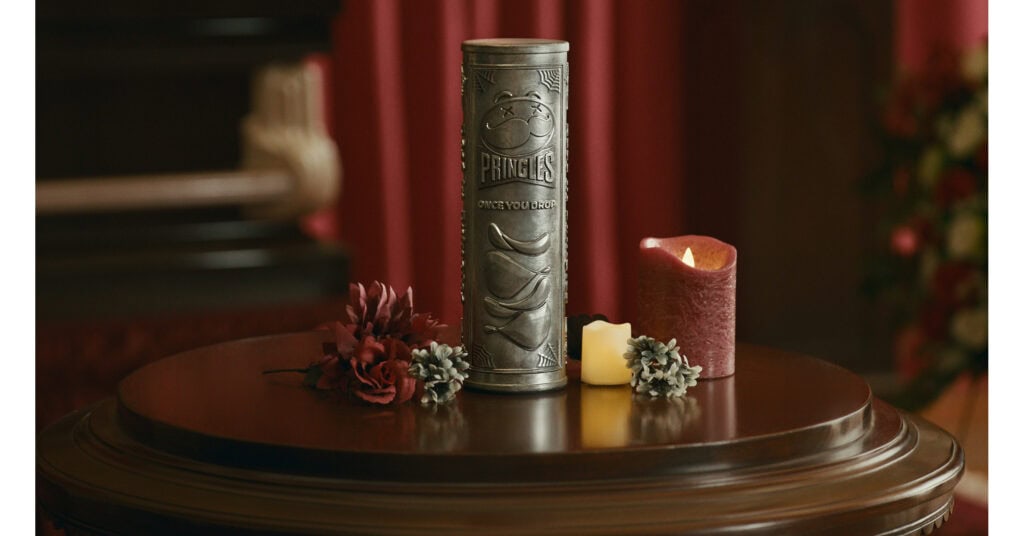 Urna prateada em formato de lata de Pringles sobre uma mesa de madeira, ao lado de flores e velas