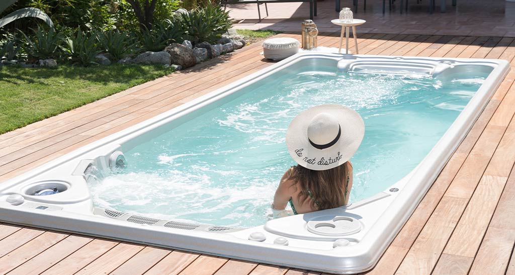 Femme de dos avec un chapeau dans un spa de nage 19EX encastré dans une terrasse en bois avec un peu de verdure sur le côté.