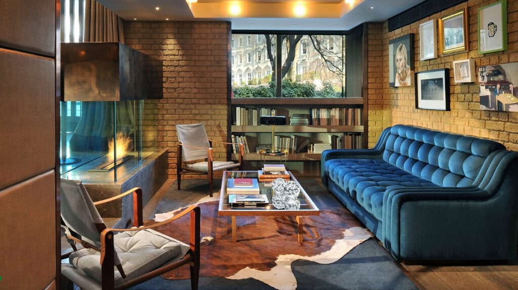 A cozy, modern living room with a blue velvet sofa, two gray armchairs, a glass coffee table on a cowhide rug, a bookshelf, framed Hari Art Prize wall art, and a fireplace, with a large window showing trees and buildings outside.