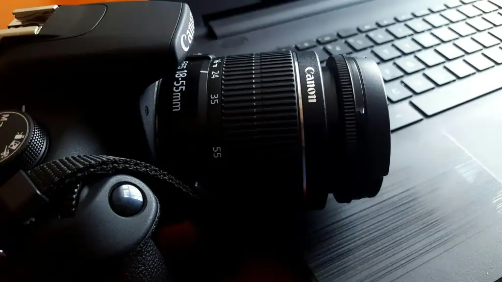 A Canon DSLR camera with an 18-55mm kit lens points to the right, placed on top of a laptop keyboard. The image is largely black and grey.