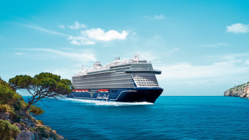 das kreuzfahrtschiff mein schiff relax fährt auf ruhigem wasser in der nähe einer küste mit bäumen und felsen im hintergrund.
