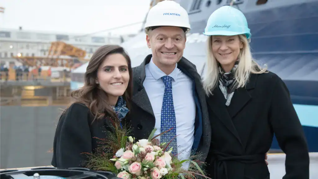 unbenannt - Michis Kreuzfahrten drei personen stehen lächelnd vor dem neugebauten kreuzfahrtschiff mit einem teil des werftgeländes im hintergrund.