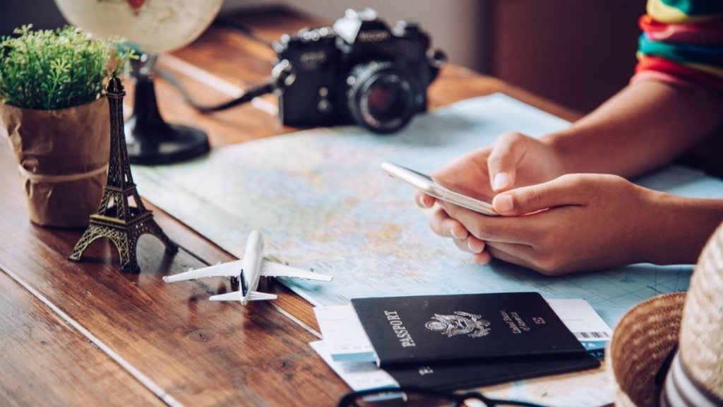 A travel-themed scene featuring a person holding a smartphone over a map on a wooden table. Nearby are travel essentials, including a U.S. passport, a small model airplane, an Eiffel Tower figurine, and a camera, evoking a sense of adventure and trip planning. 