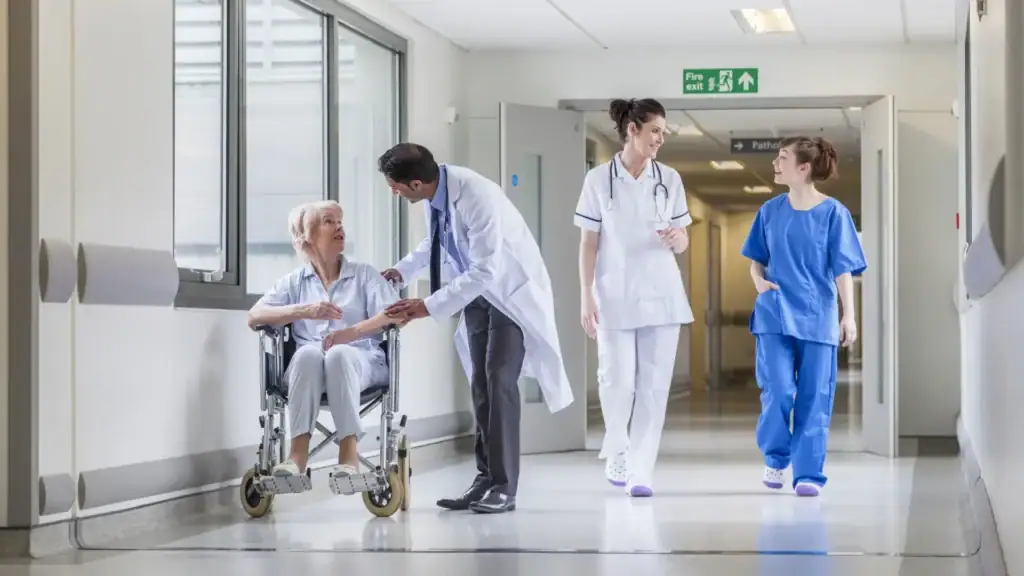 Doctor in healthcare sector assisting an elderly patient in a wheelchair while nurses walk through a hospital corridor
