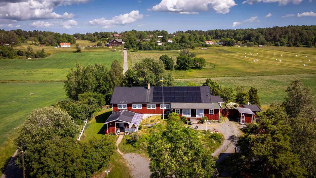 Bild på hus med solceller på taket. Flygfoto över ett rött hus med svarta solceller på taket, omgivet av grönska, grusväg och öppna fält under en blå himmel.