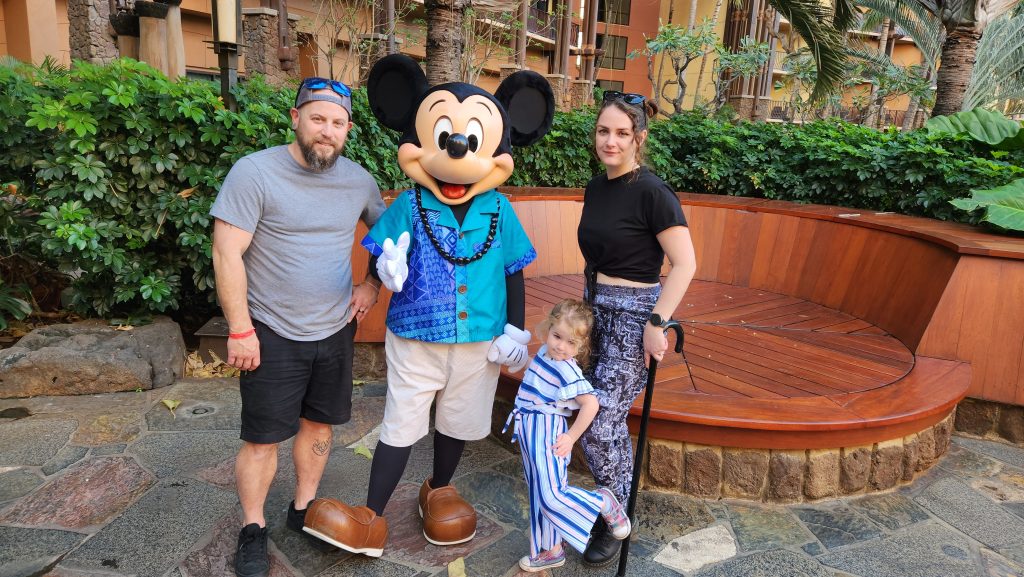 Phoenyx, her husband, and their young daughter posing with Mickey Mouse dressed in a blue Hawaiian shirt at the character breakfast in Makahiki at Disney’s Aulani Resort in Oahu. They are standing outdoors with greenery and a wooden bench behind them.