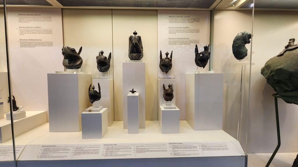 Display of ancient bronze artifacts at the Archaeological Museum of Olympia in Greece, featuring sculpted horse heads, votive offerings, and other relics arranged on white pedestals within a glass case, accompanied by descriptive plaques.