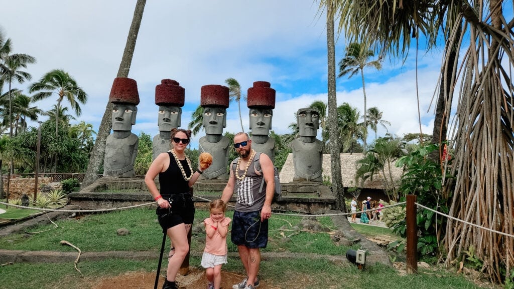A family of three poses in front of large moai statues wearing red pukao hats, surrounded by swaying palm trees and lush greenery, capturing the diverse cultures of the Pacific Islands.