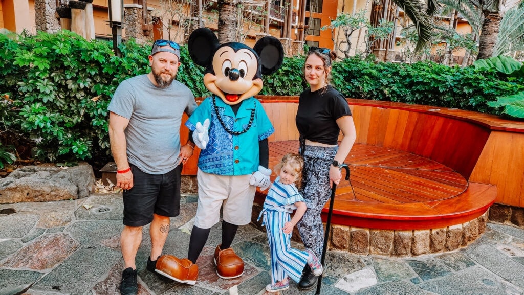 A family of three smiles for a group photo with Mickey Mouse at Aulani Resort, with Mickey dressed in a Hawaiian shirt and the little girl proudly front and center.