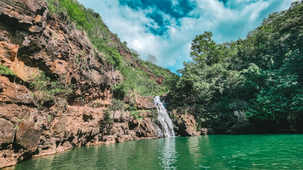 A gentle waterfall flows down a rocky cliff into an emerald green pool, framed by red rock walls and dense tropical vegetation in Waimea Valley.