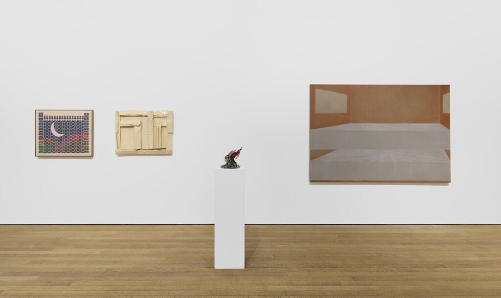 Three artworks hang on a white gallery wall at Crude Hints Towards, curated by Charlotte Edey at Ginny on Frederick for Frieze 2025. A grid moon piece, cream abstract relief, and minimalist bed painting are joined by a small dark sculpture on a white pedestal.