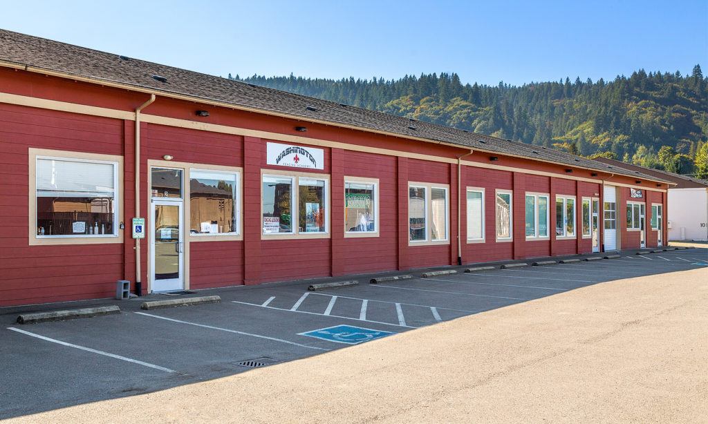 Exterior of red commercial building with multiple doors and windows, empty parking spots in front, and a forested hill in the background