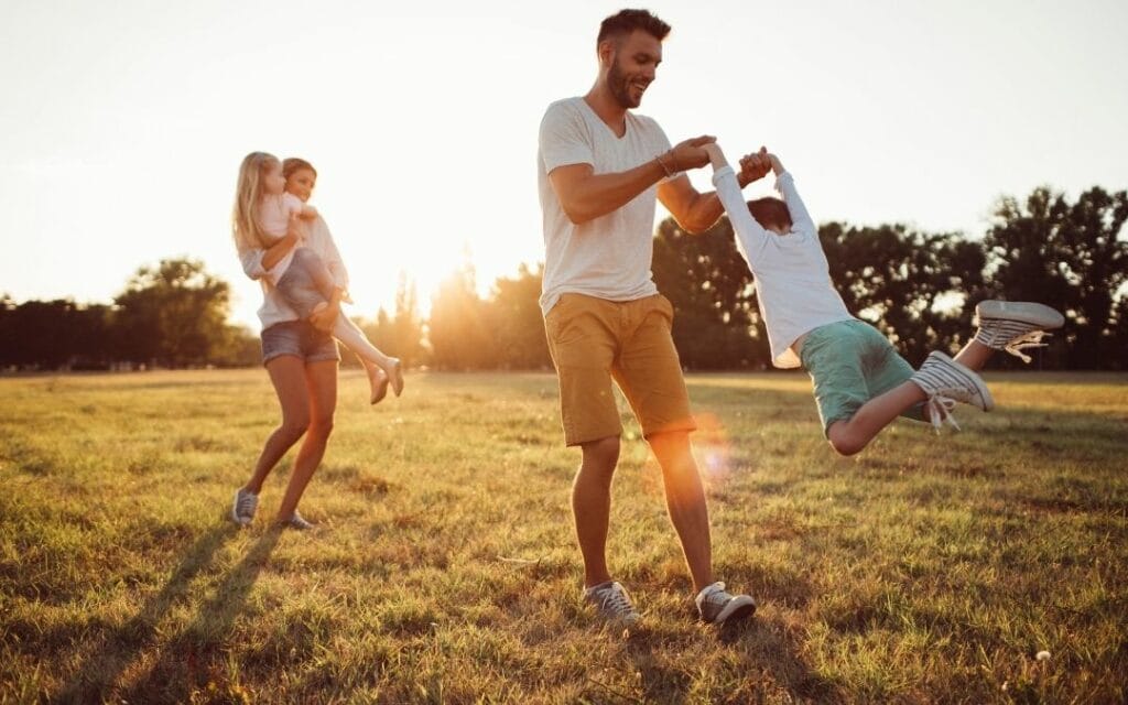 Familie mit zwei Kindern spielt draussen, die Kinder werden geschwungen und getragen: Spielen macht Oxytocin