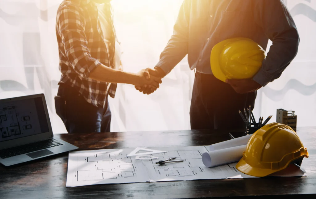 Two individuals shaking hands at a table with construction plans, indicating collaboration on a building project.