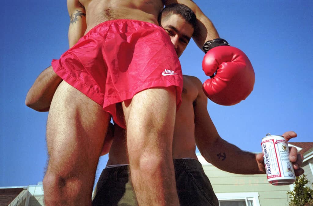 Low-angle shot of two shirtless men outdoors against a blue sky, one in red Nike shorts holding a boxing glove and a Budweiser can — photograph by Slava Mogutin