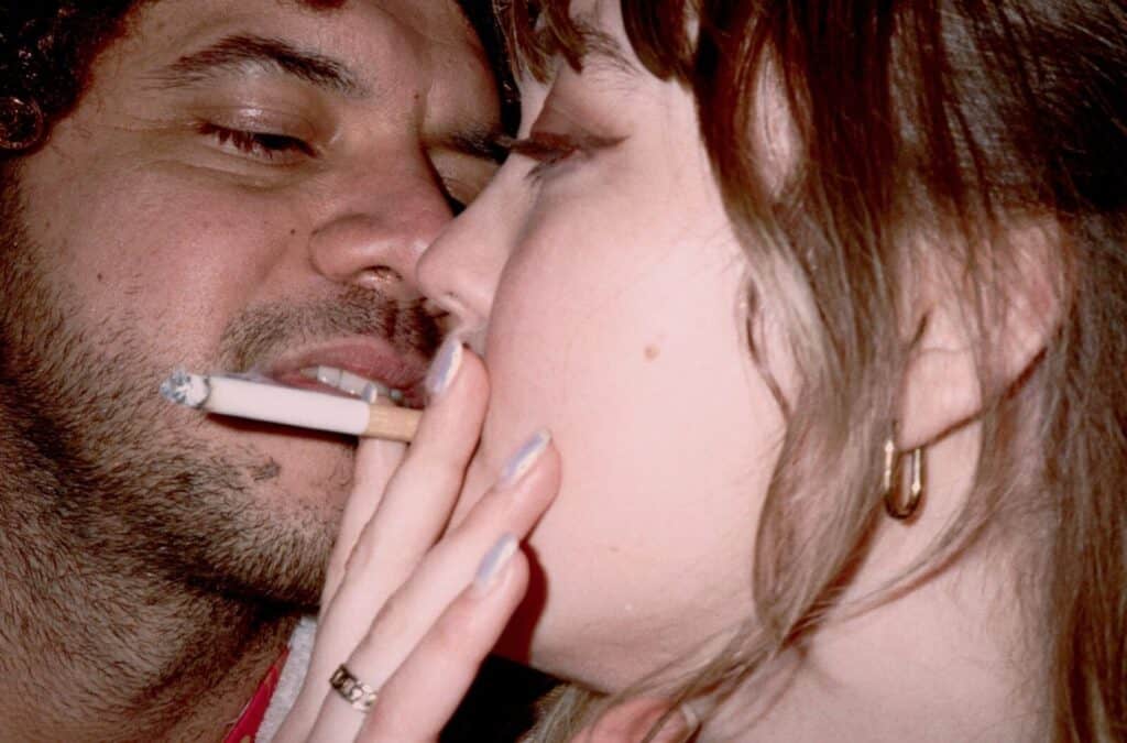 Close-up of a man and woman pressed face to face, a cigarette hanging from his lips as her hand with pale blue nails gently touches his cheek — photograph by Arthur Samier