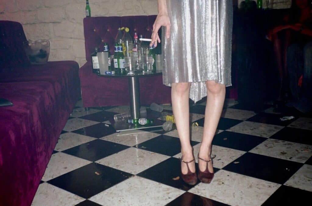 Lower half of a figure in a silver sequin midi skirt and brown T-bar heels standing on a black and white checkered floor, surrounded by fallen bottles at an after-party — photograph by Arthur Samier