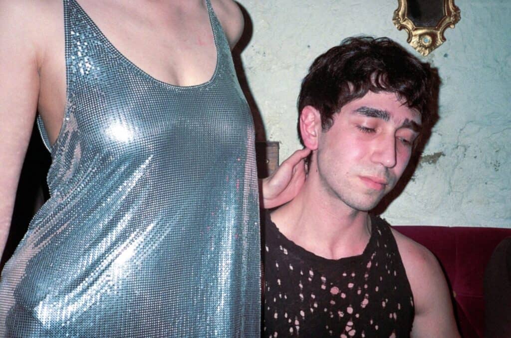 A figure in a silver chainmail dress standing beside a young man in a distressed black tank top, her hand resting on the back of his neck as he looks downward, a gilded mirror visible on the cracked wall behind — photograph by Arthur Samier