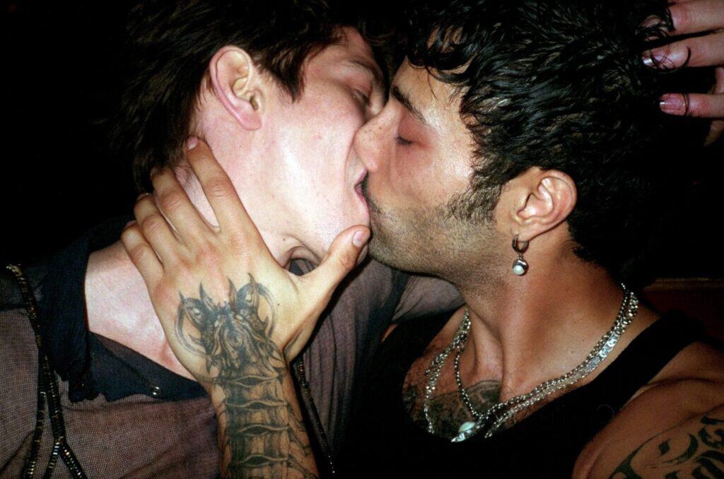 Two tattooed men kissing in a dark venue, one gripping the other's face with an intricately tattooed hand, both wearing chain necklaces — photograph by Arthur Samier