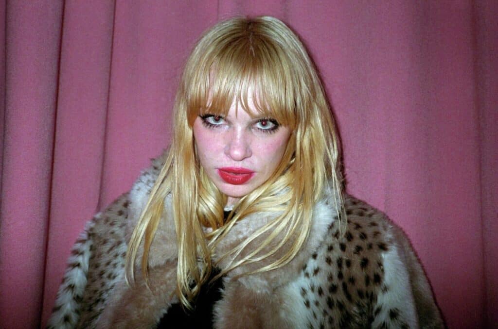 Portrait of a blonde woman with a blunt fringe, bold red lips and heavy lashes staring directly into the camera, wrapped in a leopard-print fur coat against a dusty pink curtain — photograph by Arthur Samier