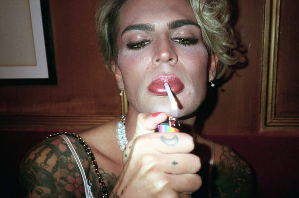 Close-up portrait of a heavily made-up figure with glossy red lips and dramatic eye makeup lighting a cigarette with a rainbow lighter, pearl necklace and gold chain visible, tattoos covering both arms — photograph by Arthur Samier