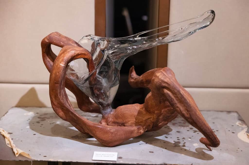 A modern sculpture featuring twisted, polished wood intertwined with a smooth, flowing clear glass element is displayed on a gray surface with a neutral background; a piece reminiscent of works seen at The Hari Art Prize Celebrating London’s Emerging Artists.