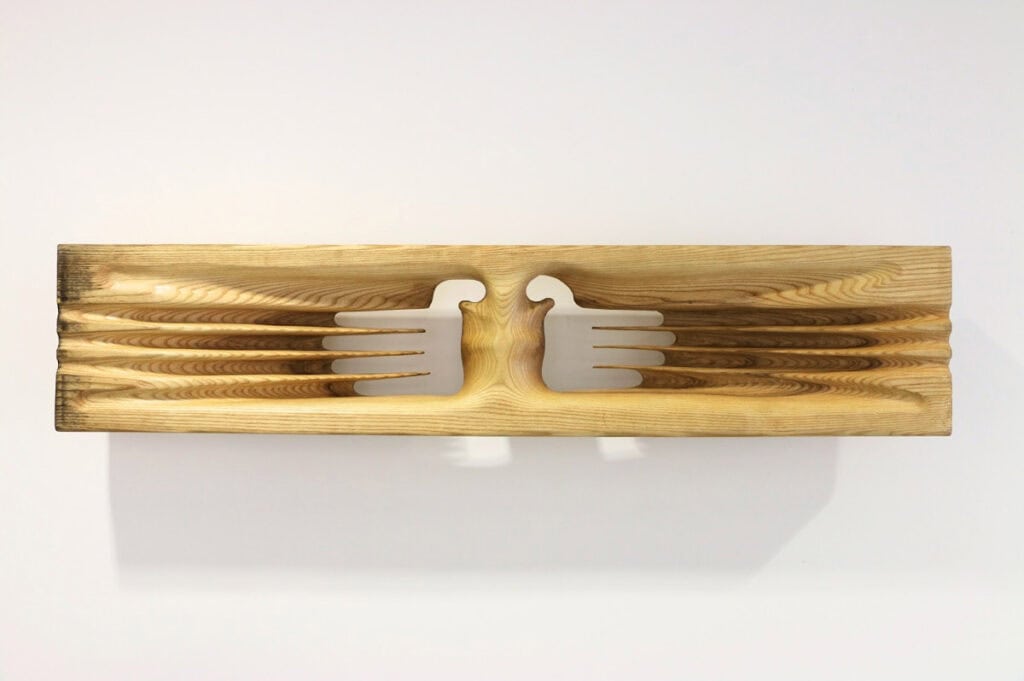 A wooden plank with two stylized bird shapes carved out, facing each other. Inspired by The Hari Art Prize’s celebration of London’s emerging artists, the wings form parallel grooves that extend outward along the wood grain in a symmetrical pattern.