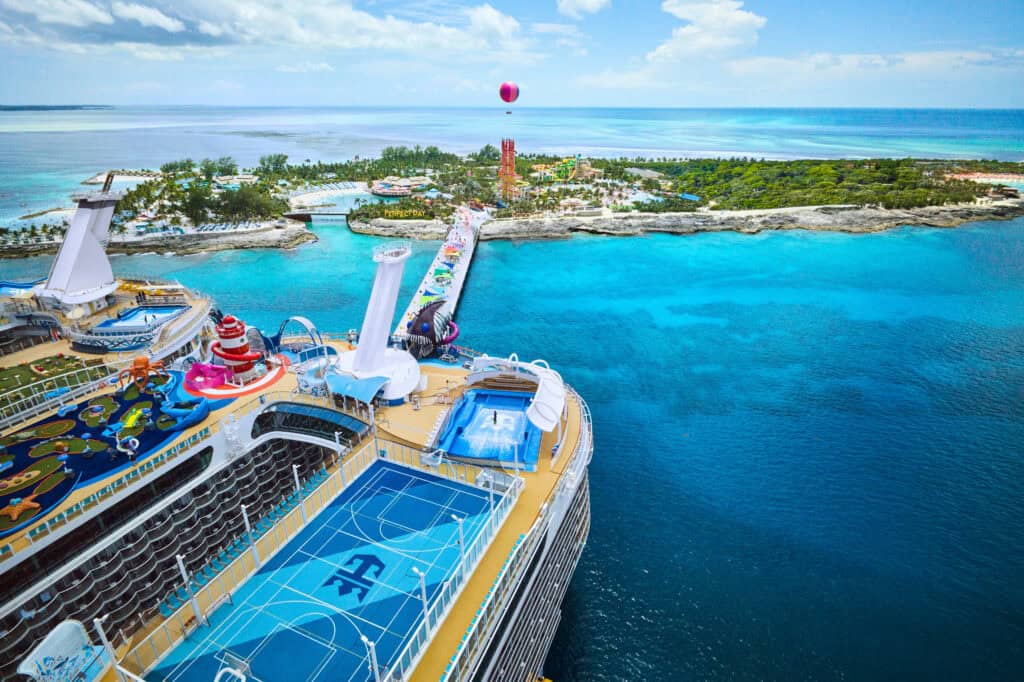 Aerial view of a cruise ship with water slides, pools, and courts near a tropical island resort.