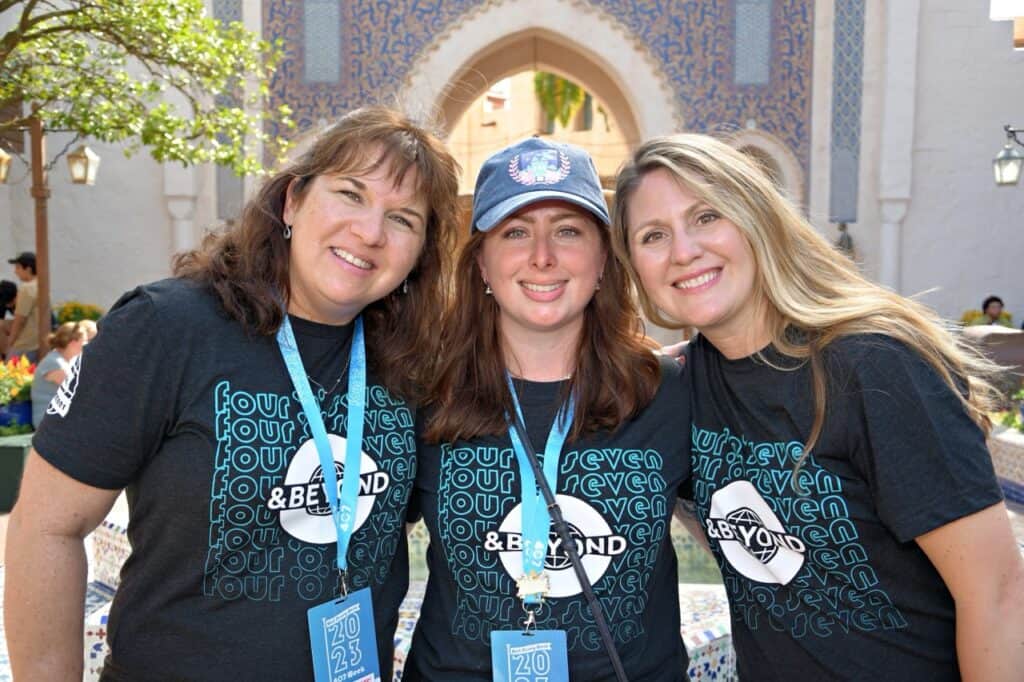 Photo of three women smiling at an outdoor event, wearing matching "seven tour & beyond" t-shirts.