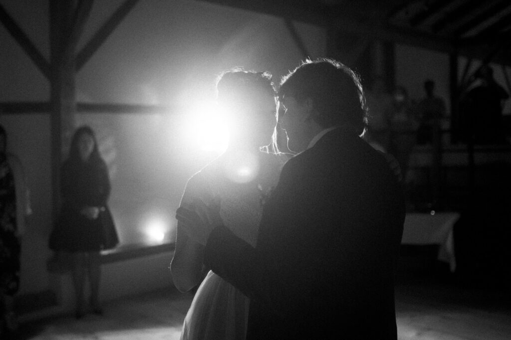 In einem schwach beleuchteten Raum tanzt ein Paar im sanften Schein einer hellen Lampe und teilt einen intimen Moment. Im Hintergrund sind Silhouetten von Figuren zu sehen, die die Magie verkörpern, die der Hochzeitsfotograf Regensburg eingefangen hat.