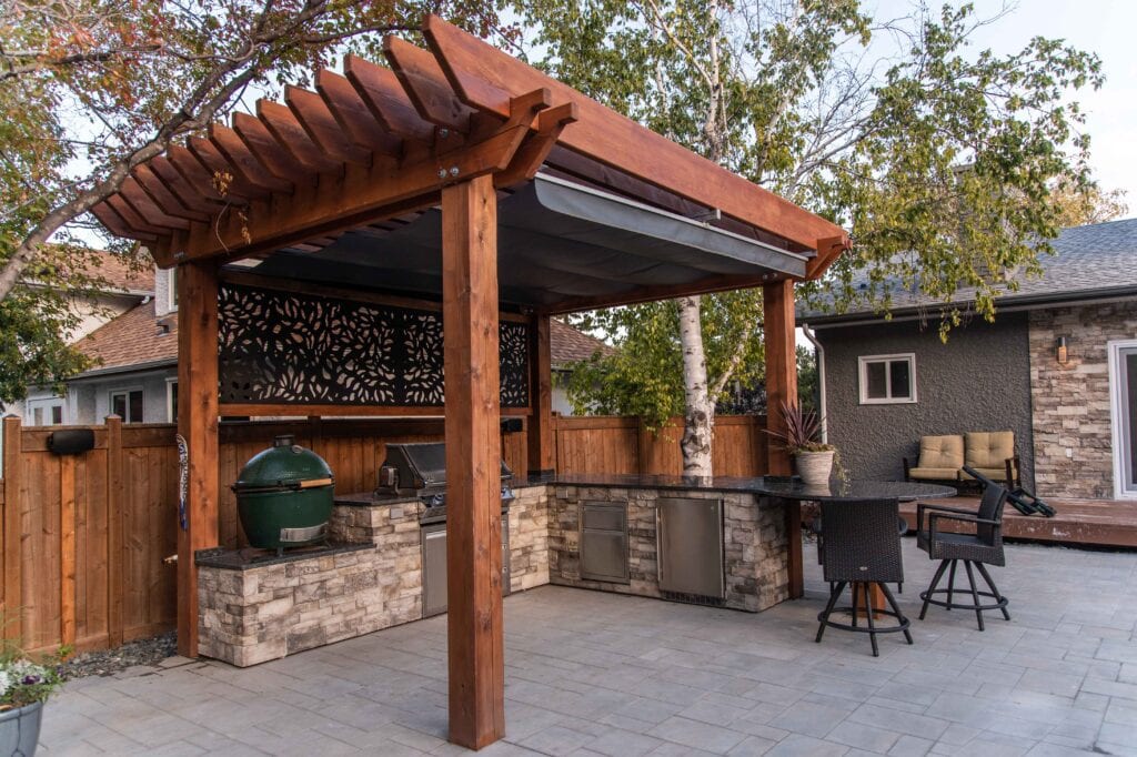 Outdoor kitchen and fireplace under pergola with stone counters and backyard landscaping.