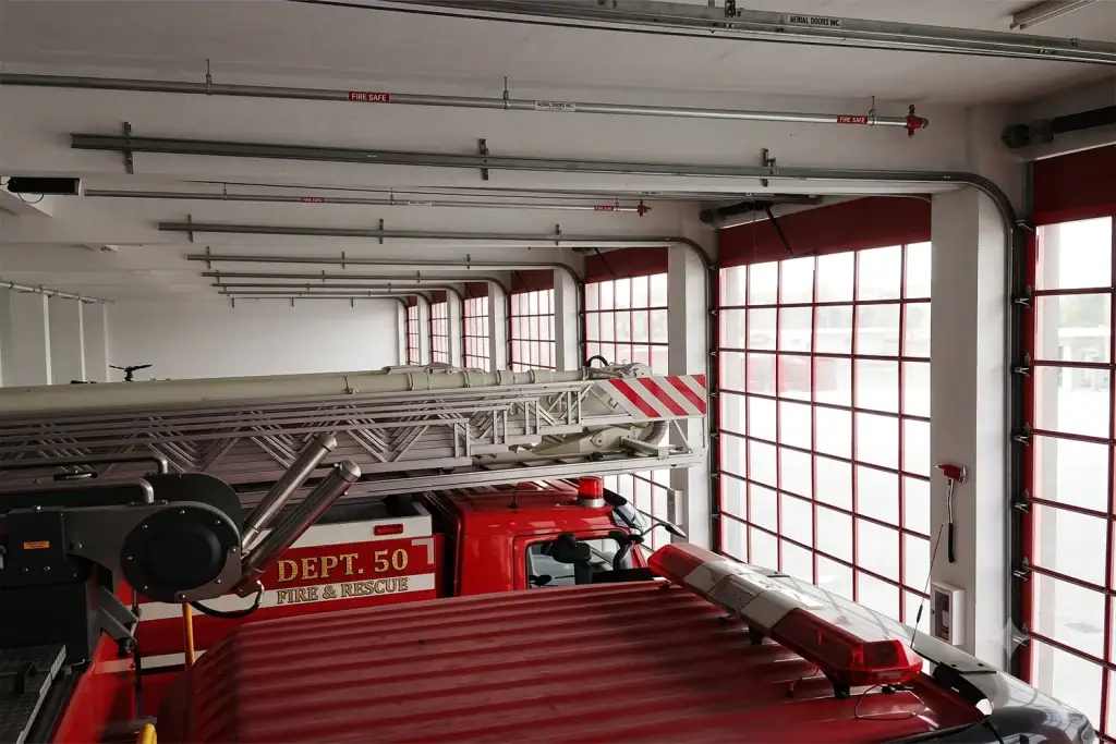 Interior view of an emergency fire and rescue station featuring commercial full-view sectional doors with red aluminum frames, showcasing the overhead track system engineered for rapid deployment and maximum visual clearance.