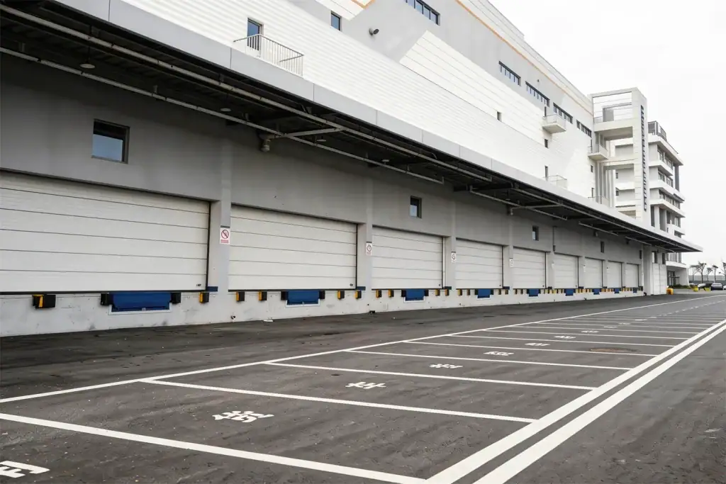 Exterior view of a massive modern logistics center featuring a continuous row of heavy-duty industrial sectional doors and blue dock levelers, demonstrating the high-volume manufacturing and delivery capacity required for mega projects.