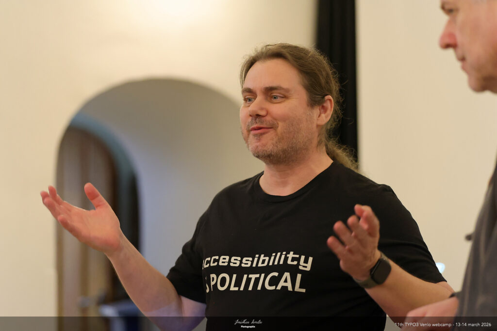 Auf seinem schwarzen T-Shirt steht "accessibility is political" (Barrierefreiheit ist politisch) - A happy Marc, smiling and talking with his hands widely spread