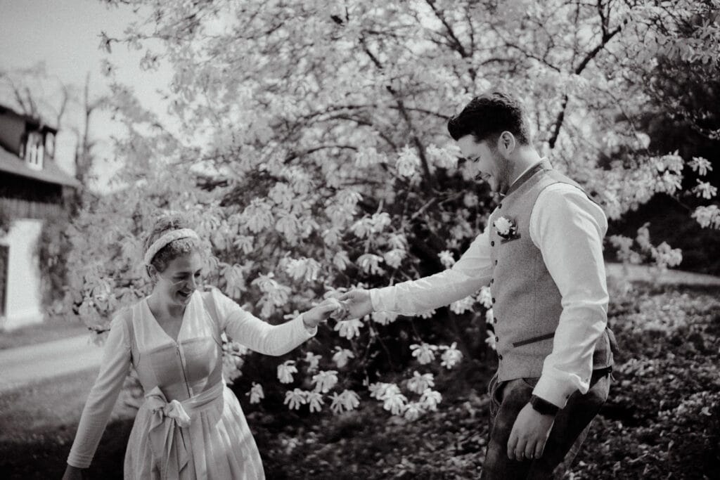 Ein Schwarz-Weiß-Bild, aufgenommen von einem Hochzeitsfotografen in Regensburg, zeigt ein fröhlich tanzendes Paar im Freien. Die lächelnde Frau in einem hellen Kleid und einem Stirnband wird anmutig von dem Mann in Weste und Hemd geführt. Sie sind von blühenden Bäumen umgeben.