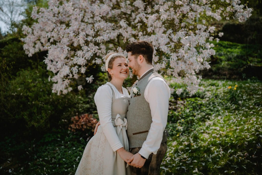 Ein Paar steht in einem üppigen Garten, hält sich an den Händen und lächelt sich an. Aufgenommen von einem Hochzeitsfotografen aus Regensburg sind sie in Hochzeitskleidung – die Braut trägt ein weißes Kleid und der Bräutigam eine Weste. Ein blühender Baum mit rosa Blüten wertet die romantische Kulisse auf.