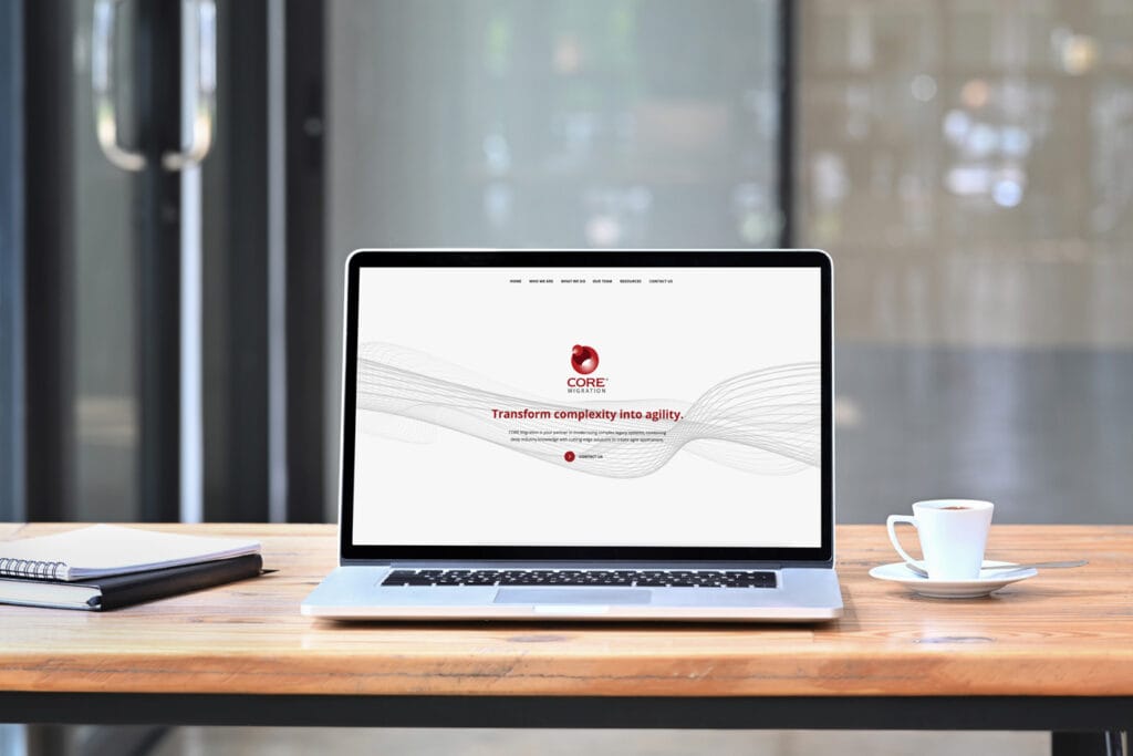 Notebook, laptop, and coffee on a table on a wooden desk in front of a window. The CORE Migration home page is visible on the laptop.