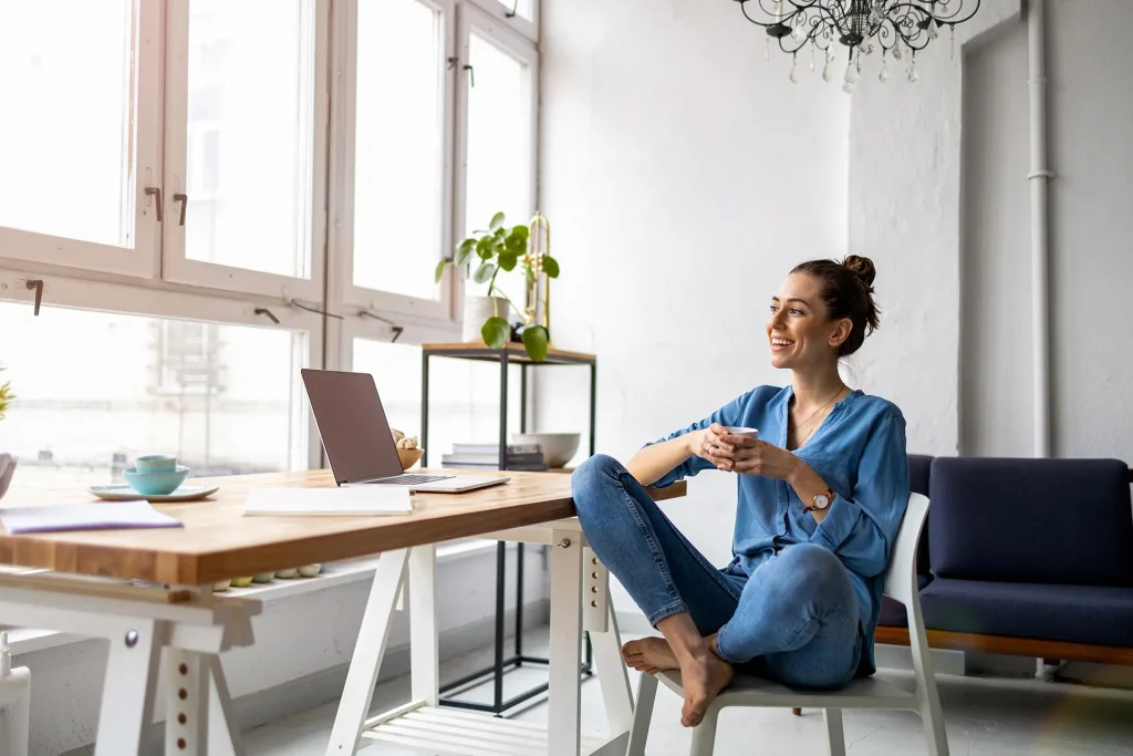 A woman sitting at a desk in a home office, smiling and holding a coffee cup, with a laptop in front of her and a window in the background, suggesting a relaxed and mindful work environment.