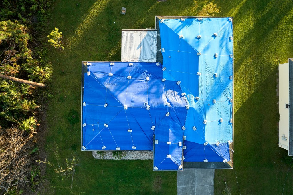 Aerial view of a house with its roof covered in blue tarps held down by sandbags, possibly after storm damage. Ledgewood Roofing & Construction provides expert roof protection and repair amid lush green grass and surrounding trees.