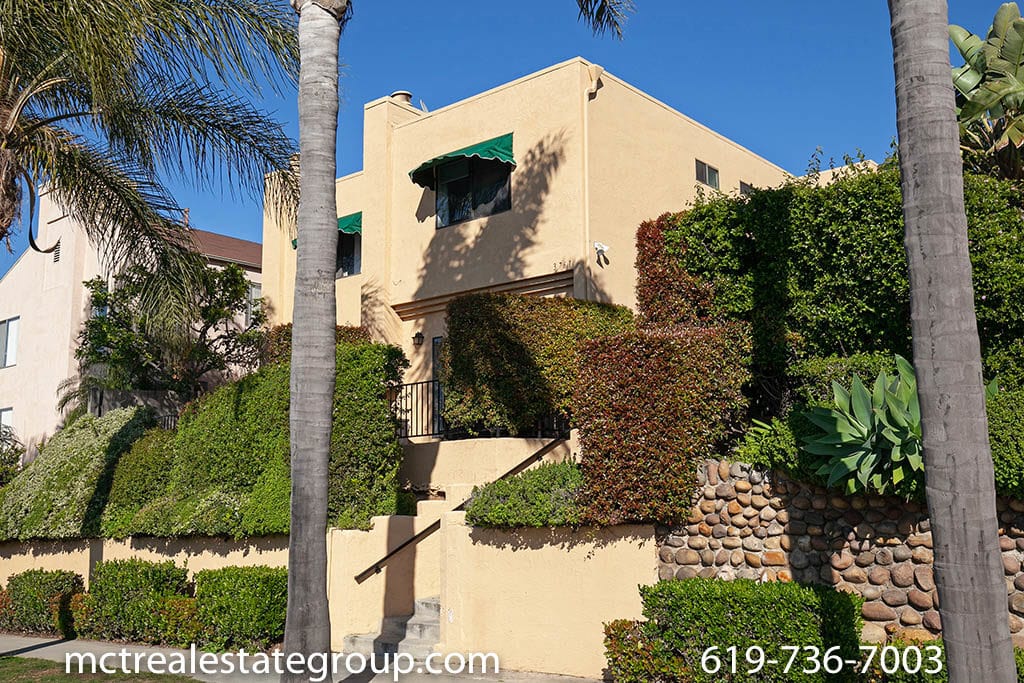 Exterior view of 3761 Villa Terrace in North Park featuring Mediterranean-style architecture with warm tan stucco, green awnings, and lush hillside landscaping accented by a decorative stone wall and towering palm trees.
