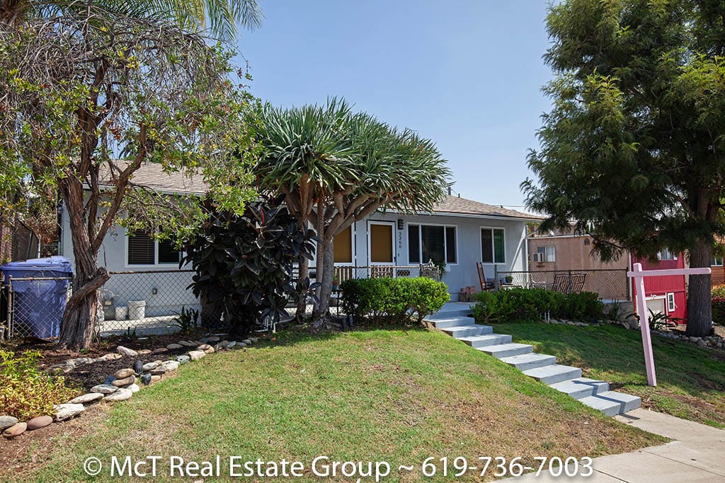 Front exterior of a white duplex at 3364-66 A Street in South Park featuring light blue concrete stairs leading up a grassy slope with tropical landscaping and a large dragon tree.