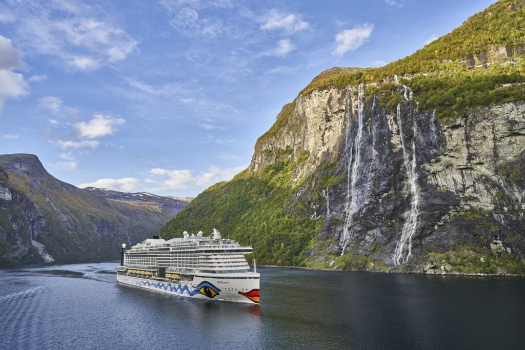 AIDA_Sommer25_Geirangerfjord_c_Skynamic - Michis Kreuzfahrten ein kreuzfahrtschiff fährt durch einen fjord, umgeben von hohen bergen und einem wasserfall im hintergrund.