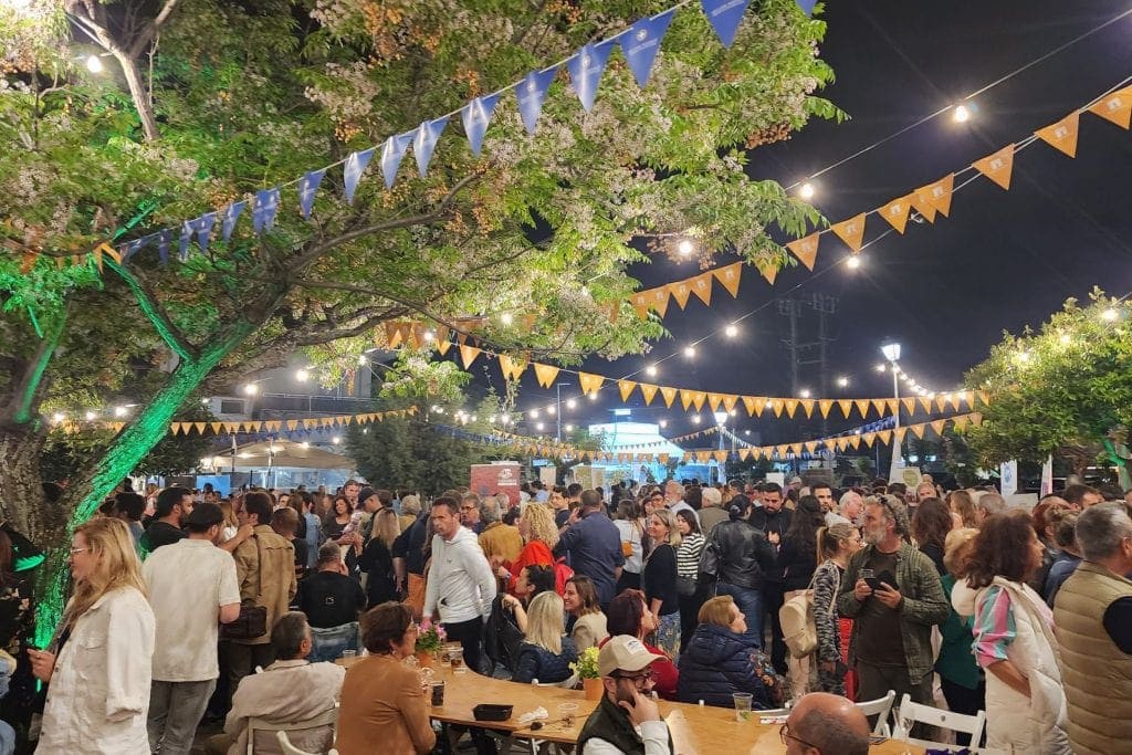 A lively Greek festival in the Kalamata city center at night, celebrating the opening party for TBEX Europe. The image features a crowd of people mingling under string lights and colorful bunting, surrounded by trees and festive decorations. 