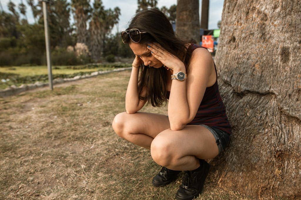 A woman kneels by a tree holding her head, expressing stress or anxiety over travel in an outdoor park. This image is also the featured image for the blog post 'How to Deal with Noise Sensitivity'.