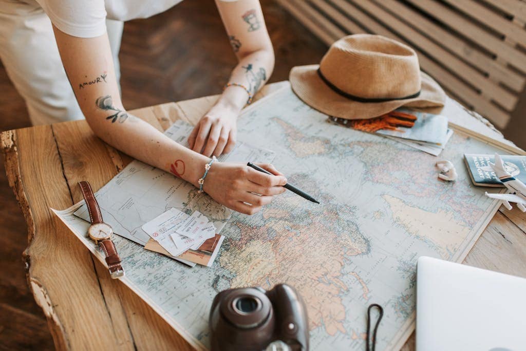 Person with tattoos leans over a large world map on a wooden table, planning a trip with a pen in hand, surrounded by travel essentials including a passport, watch, hat, instant camera, and boarding passes.