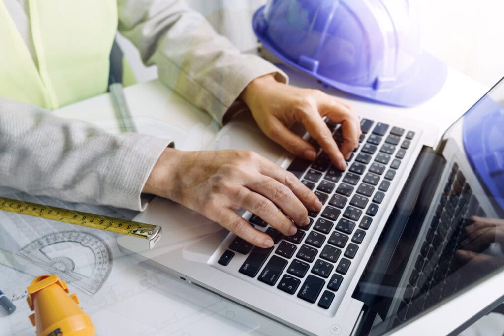 Engineer types on laptop with hard hat, ruler, and protractor, working on construction project plans.