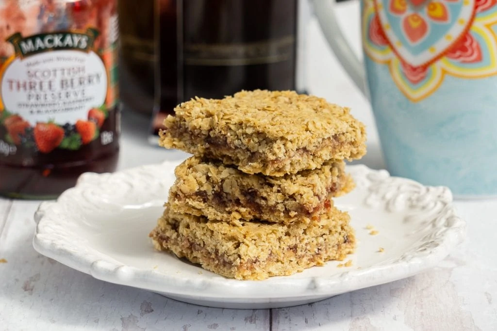 Jam Flapjacks stacked on a plate with jam jar, coffee behind