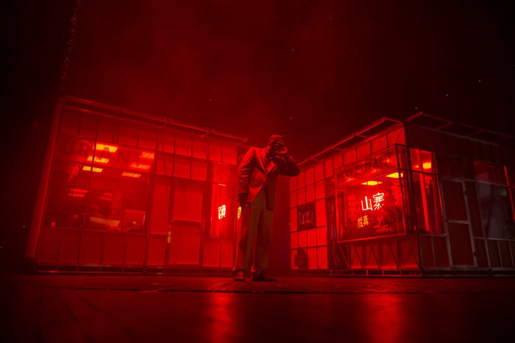 Actor standing outside under red lighting beside a building facade in a cinematic still from “ROHTKO”, photographed by Artūrs Pavlovs for Cold Magazine feature on the Barbican Theatre production.