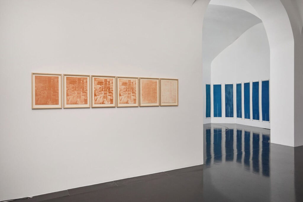 A modern art gallery features six framed orange-toned prints on a white wall; nearby, several tall blue panels from the Atto 2/3: Col Tempo exhibition curve along the shiny dark floor under high, arched white ceilings.