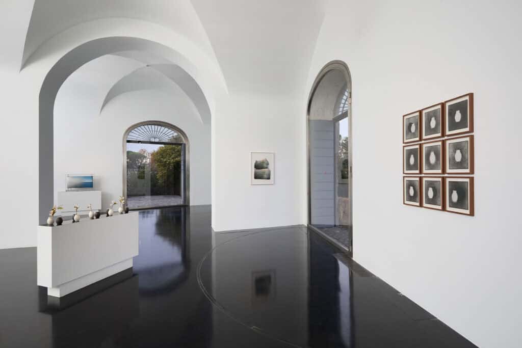 A contemporary art gallery with arched ceilings, glossy black floors, sculptures on white pedestals, and framed photos on white walls sets the stage for the Atto 2/3: Col Tempo exhibition, with natural light streaming through large arched doorways.